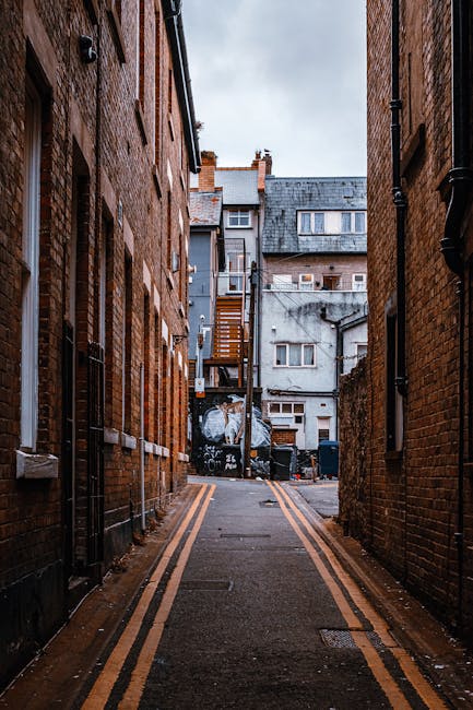 A narrow urban street between two brick buildings, showing a paved alleyway with double yellow lines along the edges. At the end of the alley, there is a building with a grey facade, a wooden staircase, and a metal fire escape. The scene includes various objects related to house removals and moving services, such as black trash bins, plastic-wrapped furniture, and cardboard boxes stacked near the building entrance. The alley is well-lit with natural daylight, and the environment appears to be in the midst of a home relocation process. Visible equipment used for furniture transport, such as trolleys and straps, suggests ongoing packing and loading activities, supporting the context of professional house removals in Kingston during a move or renovation, as managed by Movers Kingston.