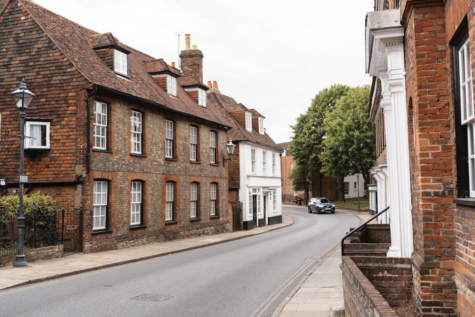 A quiet residential street in Kingston featuring a narrow road with a gentle curve. On the left, there is a row of historic brick houses with tiled roofs, multiple chimneys, and white-framed sash windows, some with small front gardens enclosed by low brick or wrought iron fences. On the right, part of a similar brick building with white decorative trim around the door and windows is visible, with a short staircase leading up to its entrance. A black vintage-style street lamp is positioned near the sidewalk on the left, and a modern grey car is seen in the distance driving along the road. The pavement is clean with a narrow curb, and a large leafy tree with green foliage extends over the street, providing some shade. The scene appears to be during daytime with diffused light under an overcast sky, capturing a typical scene suitable for home relocation or furniture transport services, as would be coordinated by Movers Kingston during a house move in Kingston KT1 with narrow street access.