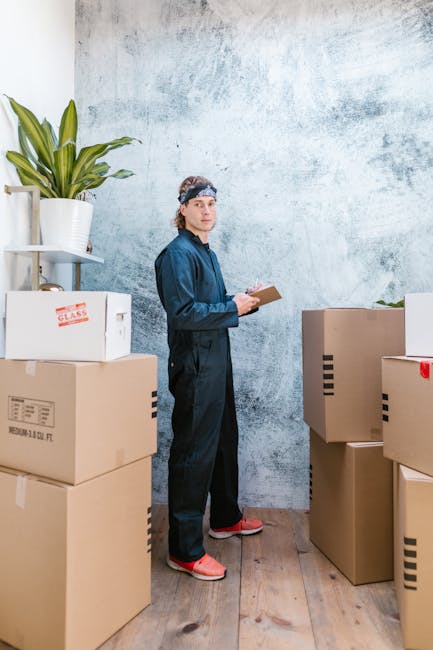 A young man dressed in dark blue work overalls and a headband stands in a room with a textured, light grey wall. He is holding a clipboard and appears to be inspecting or taking notes during a home relocation process. Surrounding him are several large cardboard boxes, some labeled with red and black markings, stacked on a wooden floor. To his left, there is a tall white shelving unit holding a green houseplant in a white pot, and some smaller items. The boxes likely contain household items being prepared for packing or transport. The scene suggests an active loading or packing environment as part of furniture transport or packing and moving services provided by Movers Kingston. The lighting is natural, and the space appears organized, with boxes positioned for easy movement and access, reflecting typical logistics involved in professional removals.