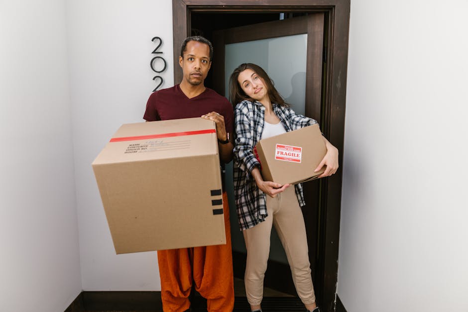 A man and a woman stand side by side in a doorway, each holding cardboard boxes during a home relocation. The man, wearing a maroon t-shirt and orange trousers, holds a large box with red and black packing tape. The woman, dressed in a plaid shirt and beige trousers, carries a smaller box marked with a 'Fragile' label, indicating the careful handling of delicate items. The doorway behind them is open, revealing a glimpse of the interior, with a dark wooden doorframe and a white wall. Inside the room, some boxes and packing materials may be visible, consistent with the packing and moving process involved in professional removals. The scene captures the active loading or unloading phase typical of furniture transport, part of a comprehensive move managed by Movers Kingston, demonstrating their approach to accurate and careful packing during house removals.
