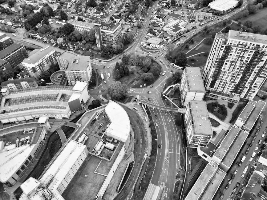 An aerial black-and-white photograph captures a busy urban area in Kingston, showing a network of roads, roundabouts, and pedestrian walkways surrounded by modern and older multi-storey buildings. In the foreground, a large building with a flat roof and various attached structures appears to be part of a commercial or office complex; nearby, there are several street-level vehicles parked along the sides of the roads. The roads include a multi-lane street with cars and trucks, with directional signage and a bridge-like pedestrian walkway crossing over the traffic. Adjacent, a curved road routes around a green space with trees, connecting to other streets leading to residential and office buildings of varying heights and architectural styles. The scene is well-lit, suggesting daytime, with visible movement of vehicles indicating active loading and unloading activities typical of house removals and furniture transport. This setting relates to the process of home relocation as managed by Movers Kingston, highlighting the urban infrastructure involved in moving and packing services in the Kingston area.