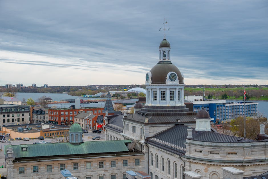 A high-angle view of Kingston town centre showing the historic borough buildings with a prominent clock tower, part of the County Hall, in the foreground. The building features ornate white stonework, a dark domed roof, and a weather vane on top. To the left, rooftops with green and slate coverings are visible, along with several multi-storey commercial and residential structures. In the background, a large body of water stretches across the scene, with boats docked along the shoreline and additional buildings beyond. The sky above is overcast with grey clouds, creating a muted lighting environment, suitable for illustrating the context of a home relocation or furniture transport process involving nearby historic and commercial properties, as handled by Movers Kingston, during packing and loading operations.