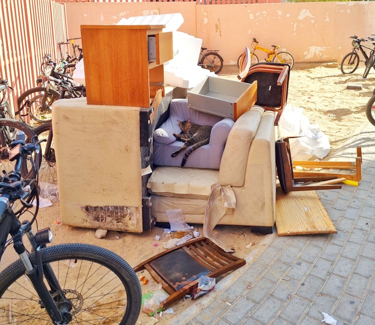 An overflowing public rubbish bin on a pavement in Kingston, surrounded by numerous black, white, and brown waste bags, cardboard boxes, and various packaging materials spilling onto the ground. The b