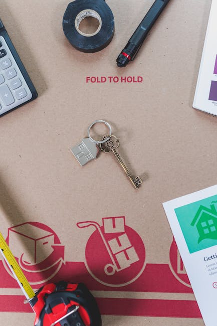 A flat-lay image showing a beige work surface used for packing and moving preparation. Visible objects include a black marker pen with a red tip, a roll of black tape, a silver key attached to a small transparent plastic keychain, and a white calculator with grey buttons. Partially visible on the edge is a yellow measuring tape, and below it, a cardboard moving box or packing paper with printed diagrams and instructions related to home relocation. The cardboard features red and green graphics illustrating packaging and furniture transport processes, alongside text instructing to 'FOLD TO HOLD.' The overall scene suggests workspace setup for furniture transport and logistics planning by movers such as Movers Kingston, supporting the theme of house removals and moving services.