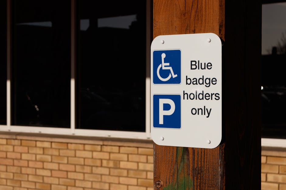 A close-up image of a white parking sign attached to a wooden post outside a building with large windows reflecting the surroundings. The sign features a blue disability badge symbol at the top, follo