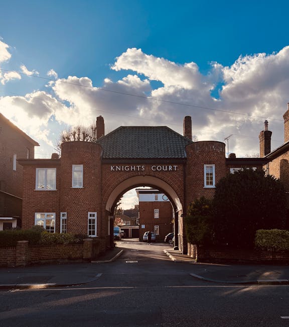 A brick residential building with an arched gateway in the center, labeled 'Knights Court' at the top, situated on Kingston High Street. The structure features small windows and two cylindrical towers on either side of the arch. Behind the building, parked cars are visible through the archway, with additional brick buildings further in the background. The sky is partly cloudy with patches of blue, and the scene is illuminated by natural daylight. In the foreground, the street and pavement are visible, with some greenery including a bush on the right side, framing the scene. The image captures the entrance to a residential area in Kingston, which may facilitate home relocation or furniture transport activities, relevant to house removals services that Movers Kingston might undertake.