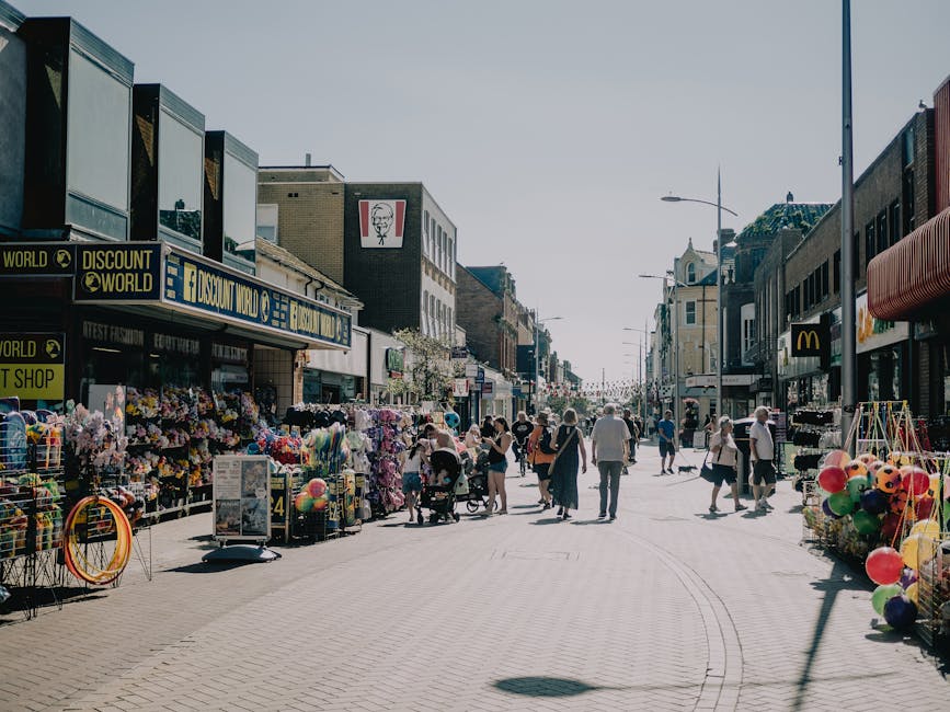 The image captures a busy shopping street during daytime, lined with various retail stores including a KFC fast food outlet and a McDonald's, with outdoor stalls on the pavement displaying an assortment of colourful goods such as flowers, balloons, and household items. Pedestrians are walking along the wide pedestrianised area, some pushing strollers or carrying shopping bags, while others browse the street vendors. Several people are engaged in lifting or carrying boxes and packages, which are wrapped in cardboard and plastic, suggesting a home relocation or packing process. Visible equipment includes hand trolleys and straps being used to transport the boxes, and parts of a moving van are visible, indicating loading or unloading activities. The scene is illuminated by natural daylight with clear skies, and the street features modern streetlights. This busy urban environment, with its variety of objects and ongoing logistical activity, reflects the typical setting for house removals and furniture transport, highlighting the crowded nature of relocation logistics.