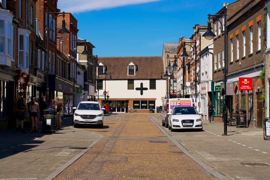 A brick residential building with an arched gateway in the center, labeled 'Knights Court' at the top, situated on Kingston High Street. The structure features small windows and two cylindrical towers on either side of the arch. Behind the building, parked cars are visible through the archway, with additional brick buildings further in the background. The sky is partly cloudy with patches of blue, and the scene is illuminated by natural daylight. In the foreground, the street and pavement are visible, with some greenery including a bush on the right side, framing the scene. The image captures the entrance to a residential area in Kingston, which may facilitate home relocation or furniture transport activities, relevant to house removals services that Movers Kingston might undertake.