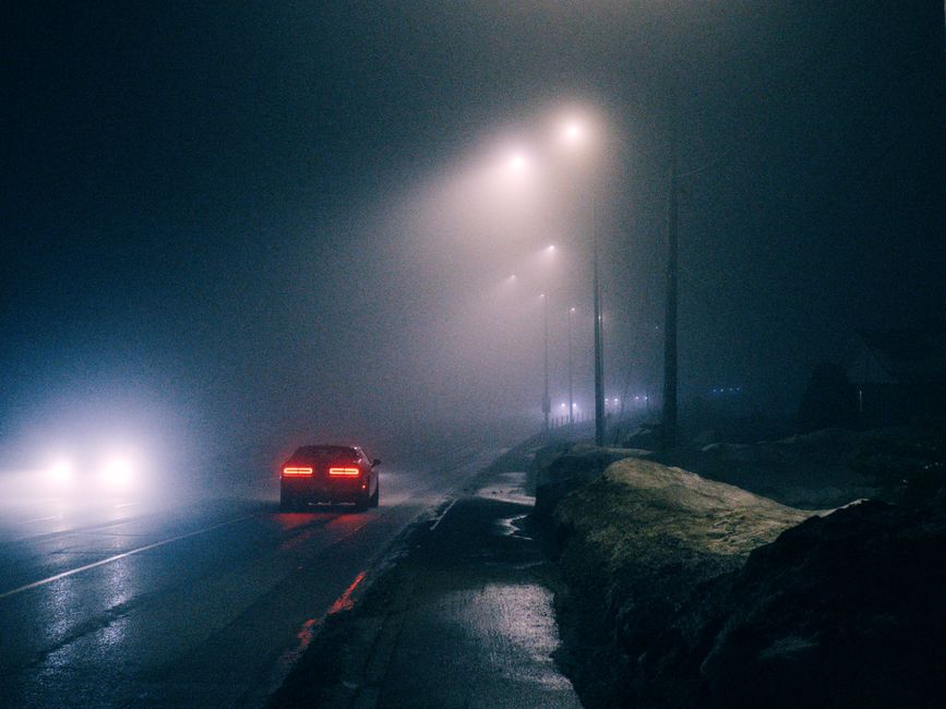 A dark, foggy night scene showing a wet road illuminated by tall streetlights with their glow diffused by the fog. A red car is parked on the side of the road, with its rear lights on, while another vehicle approaches from the distance with bright headlights cutting through the mist. To the right, a snow-covered pavement is visible alongside a pile of large rocks or snowdrifts, indicating low visibility conditions. The scene captures an outdoor environment typical of winter or early morning, with atmospheric lighting creating a subdued, muted ambiance. This image could relate to the logistical challenges of home relocation or furniture transport in low-visibility conditions, where careful planning and appropriate vehicle equipment from companies like Movers Kingston would be essential for successful removals or moving services.