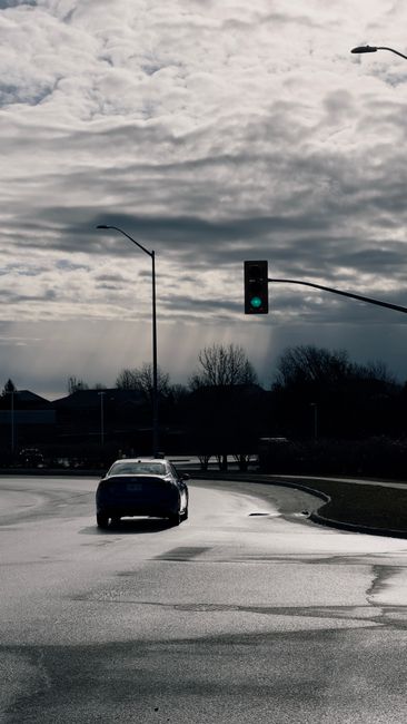 Photograph of a car waiting at a traffic light on a wet, paved street during daytime with overcast skies. In the background, there are leafless trees and low-rise buildings, with the traffic light showing a green signal to the vehicle. The image captures a typical urban street scene, relevant to home relocation and moving logistics, illustrating the need for careful planning when navigating narrow drives and low bridges during furniture transport and packing and moving processes. The scene emphasizes the importance of precise vehicle maneuvering and adherence to traffic signals during residential removal services, as provided by Movers Kingston.
