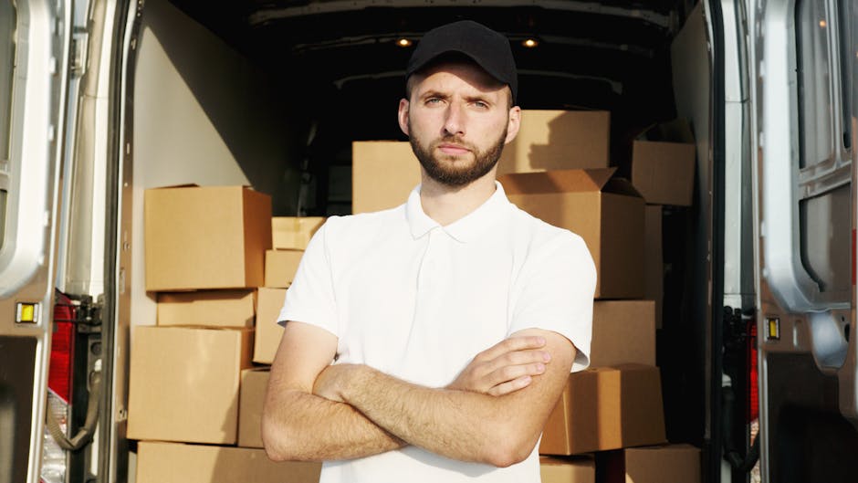 A young man with a serious expression, wearing a black cap and a white polo shirt, stands with arms crossed inside the open back of a moving van. Behind him, there are neatly stacked cardboard boxes of various sizes, some wrapped in plastic for protection during a house move. The van is parked outdoors on a paved surface, with the interior visible, showing additional boxes and packing materials. The scene depicts a home relocation and furniture transport process, highlighting the loading process managed by movers Kingston. The lighting is natural, and the image captures the preparation for a packing and moving service as part of a same-day removal in Kingston, emphasizing efficient logistics without any promotional elements.