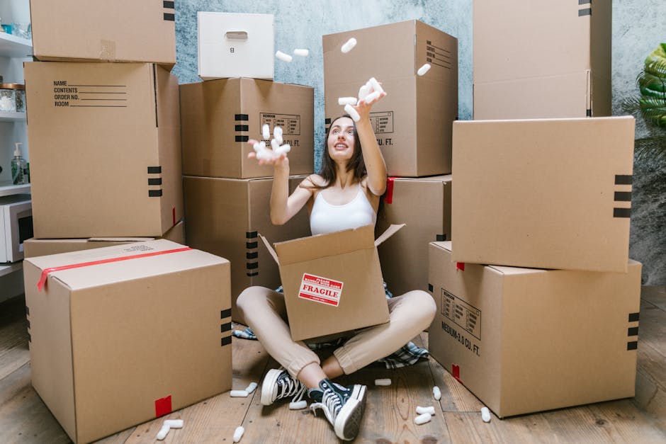 A woman sitting on the floor inside a property surrounded by numerous large cardboard moving boxes, some sealed with red packing tape and others labeled with handling instructions, as she playfully throws foam packing peanuts into the air. The boxes vary in size and are stacked against a textured blue wall, with a doorway visible behind her. She is wearing casual clothing, including a white tank top, beige pants, and black-and-white sneakers. A cardboard box with a 'Fragile' sticker is open in her lap, indicating packing and moving preparation. To her left, a small white shelf holds a few household items, and on the right, a potted plant adds a touch of greenery to the scene. The environment suggests an active house relocation or furniture transport process, with moving equipment like foam peanuts and the focus on packing materials. Image is relevant to professional removals services such as those provided by Movers Kingston, emphasizing packing and home relocation logistics.