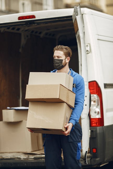 A young man with a serious expression, wearing a black cap and a white polo shirt, stands with arms crossed inside the open back of a moving van. Behind him, there are neatly stacked cardboard boxes of various sizes, some wrapped in plastic for protection during a house move. The van is parked outdoors on a paved surface, with the interior visible, showing additional boxes and packing materials. The scene depicts a home relocation and furniture transport process, highlighting the loading process managed by movers Kingston. The lighting is natural, and the image captures the preparation for a packing and moving service as part of a same-day removal in Kingston, emphasizing efficient logistics without any promotional elements.