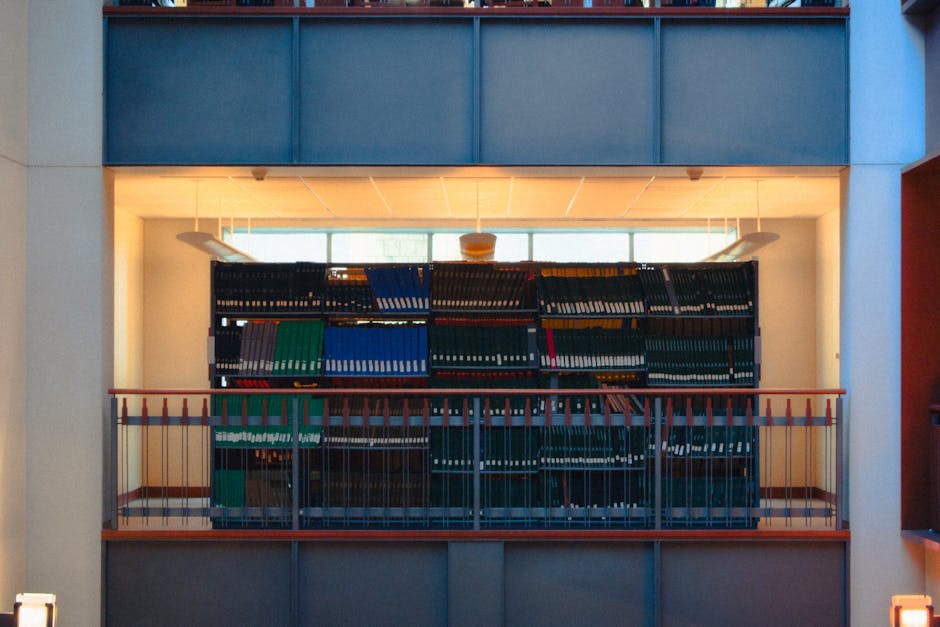 Inside a modern building with large windows allowing natural light, a black metal shelving unit is positioned against a white wall, filled with numerous closed binders and folders in various colours including green, blue, black, and grey, arranged neatly in rows. The shelving unit spans most of the width of the inner space, which appears to be a study or library area. In front of the unit, a wooden handrail with vertical slats separates the area from what looks like a balcony or elevated walkway. The floor beneath is not visible. On the right side, part of a cardboard box and packing materials are visible, suggesting a packing or moving process. The environment has an orderly, professional appearance, indicative of a setting where items are being prepared for transport or relocation, consistent with services provided by Movers Kingston for house removals. This setting likely depicts an interim storage or packing area during the furniture or document transfer involved in a home relocation from Surbiton Station to Kingston University.